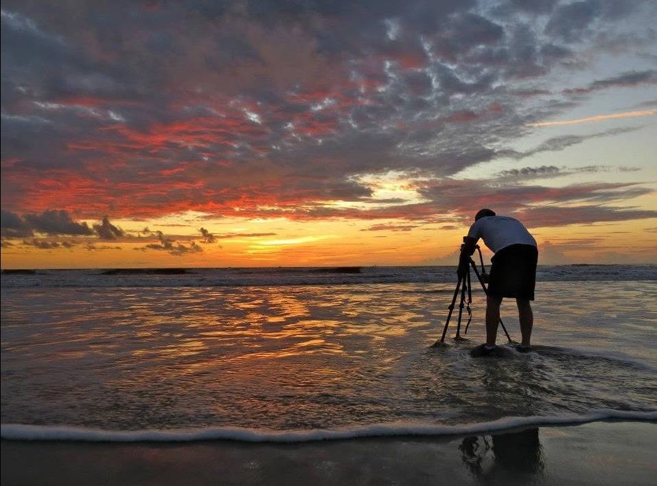 Bent over a tripod set in the surf, filming a red and orange sunset over the ocean.