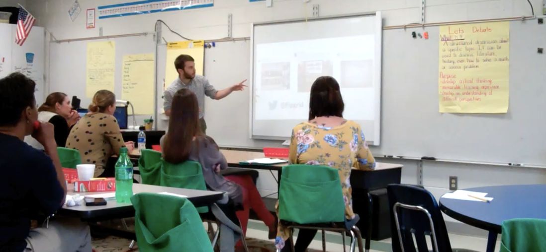 Leading a professional learning session, pointing at a projected slide for a room of teachers at classroom tables.