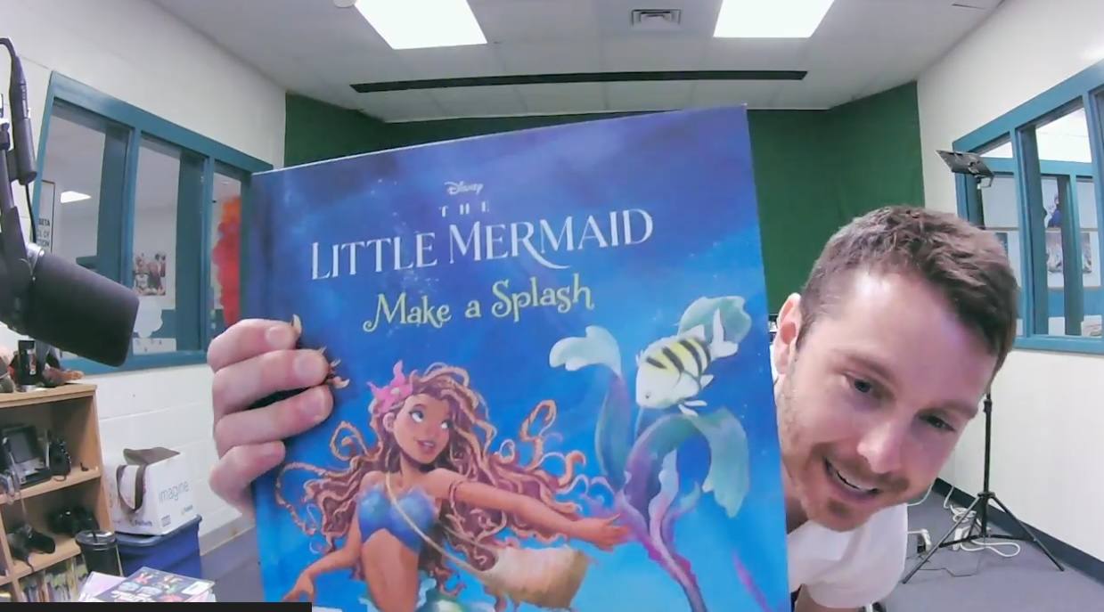 Holding a picture book up to the camera in the green-walled studio, with a microphone and light stand in frame.