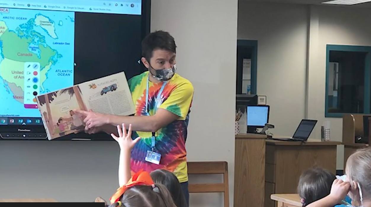 Reading a picture book aloud to a class in a tie-dye shirt, with a map of North America on the board behind and a student’s hand raised.