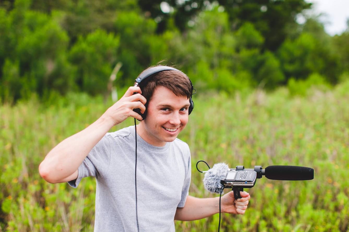 Wearing headphones and holding a field recorder with a shotgun microphone in a green field.
