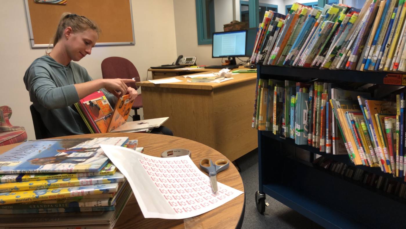 Genre stickers being applied to picture books at a round table, with a sticker sheet, book tape, and scissors laid out and a cart of color-coded spines beside it.