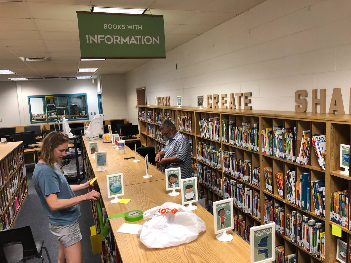 Sticky notes and framed genre signs going up along the shelf tops under the Books With Information banner, with THINK, CREATE, SHARE lettered on the wall.