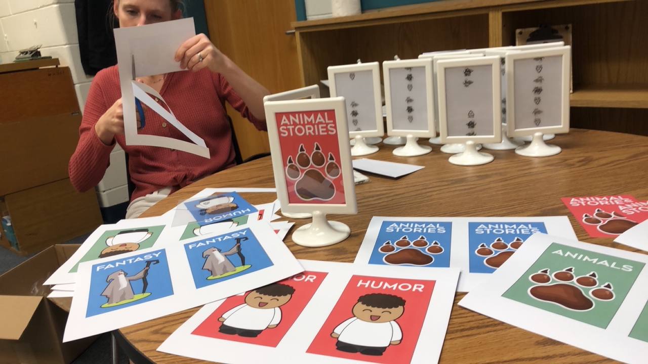 Cutting printed genre signs at the round table, with Animal Stories, Fantasy, and Humor signs in three colors and a row of empty stand-up frames behind.
