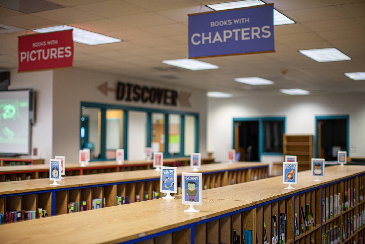 The rebuilt library: a red Books With Pictures banner and a blue Books With Chapters banner hang over shelving runs topped with framed genre signs, with a wooden DISCOVER arrow on the far wall.