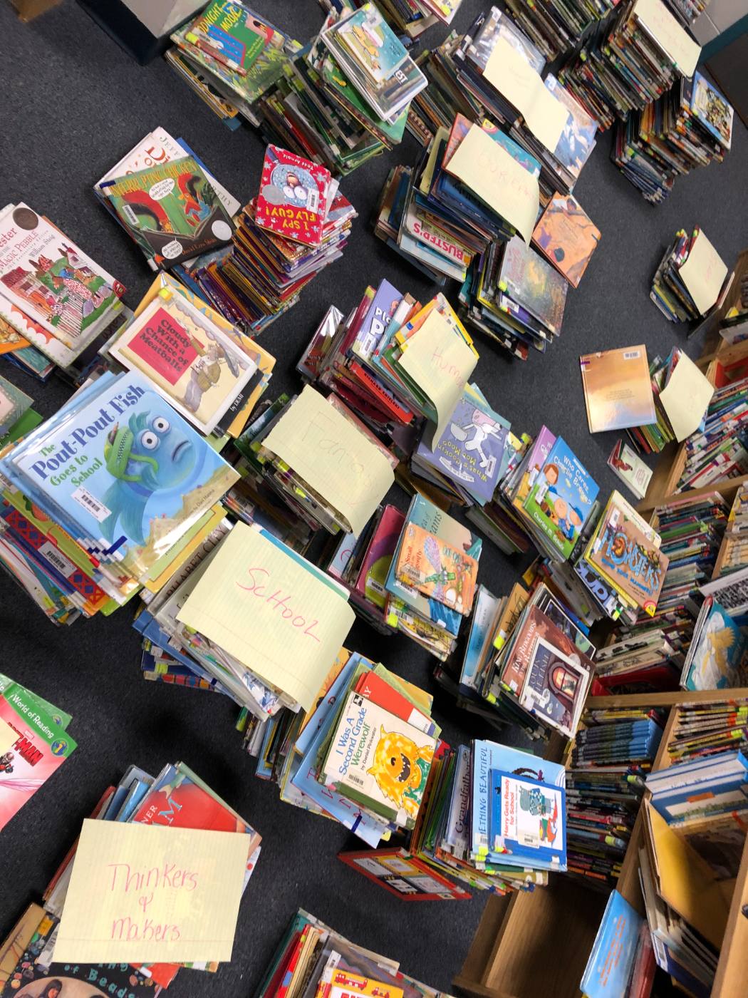 Picture books in piles across the carpet, each pile topped with a yellow sheet naming its genre: School, Fantasy, Humor, Thinkers and Makers.