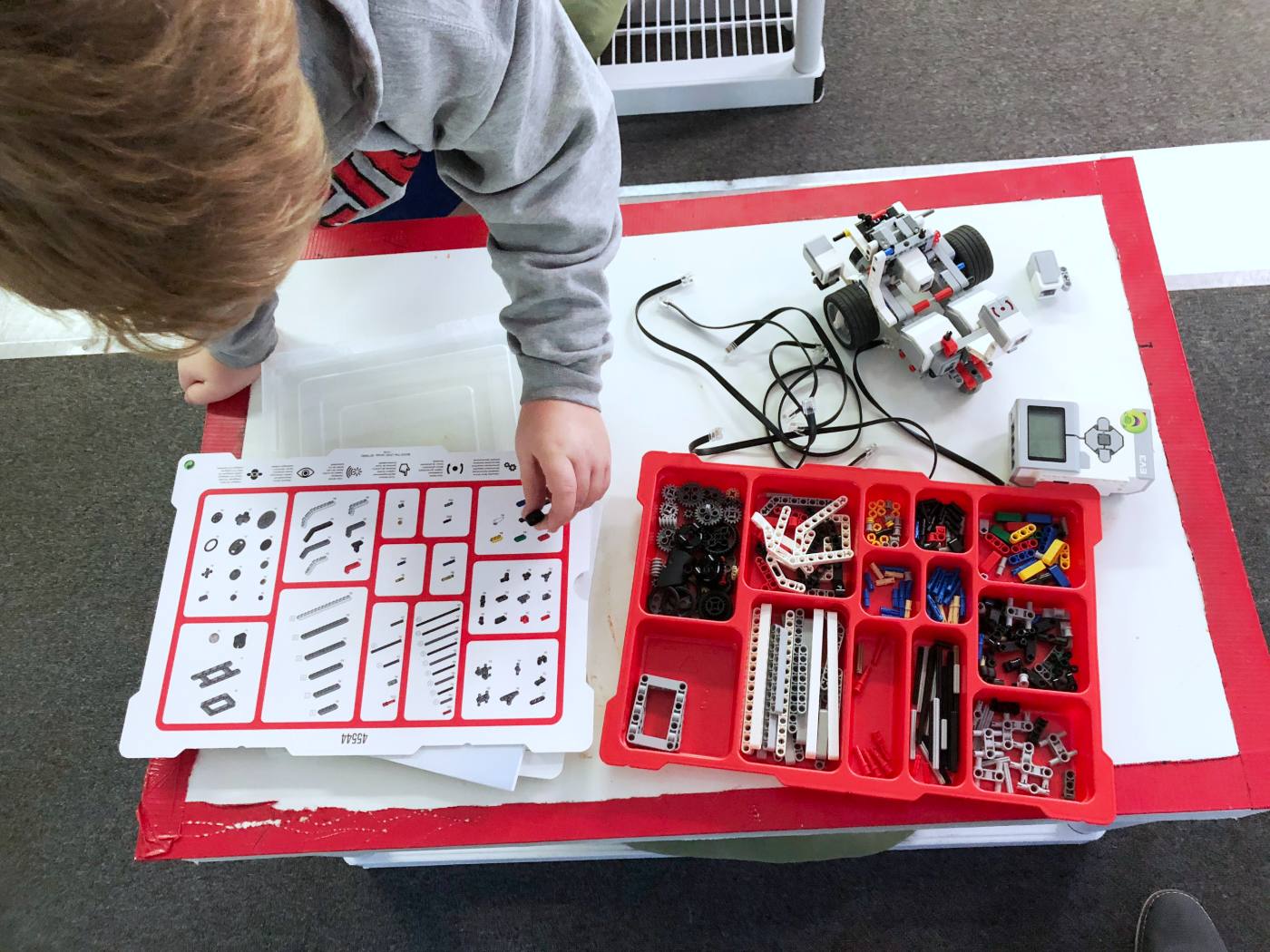 A student picks LEGO pieces from a red tray of sorted parts on a whiteboard table, with the robot and its build sheet beside it.