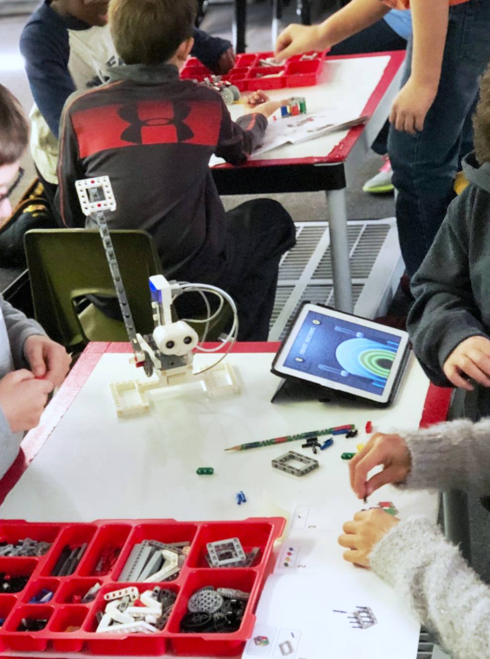 Students building and coding LEGO robots at the whiteboard tables, with parts trays and a tablet on the surface.