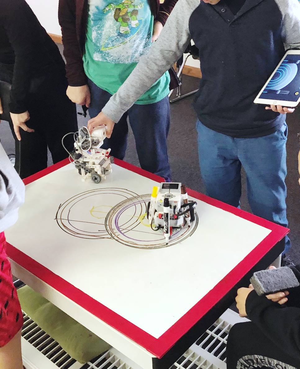 A LEGO robot carrying markers draws overlapping circles on a whiteboard table while students watch and one steers from a tablet.
