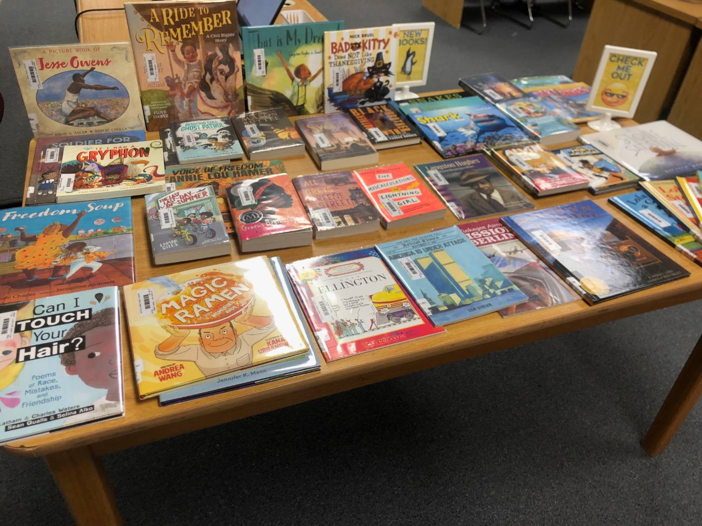 A library table covered in featured books laid flat and standing, with New Books and Check Me Out signs.