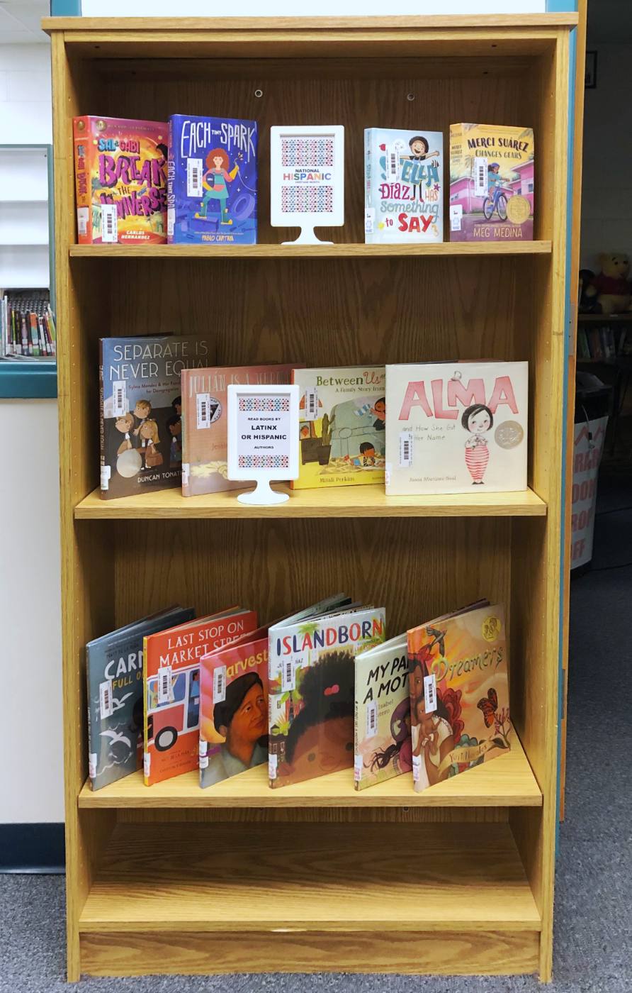 A tall bookcase of books by Latinx and Hispanic authors, with framed National Hispanic Heritage Month signs on two shelves.