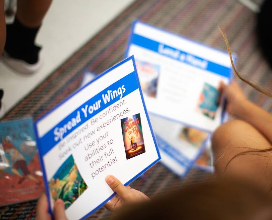 A student holds a Spread Your Wings theme card with book suggestions, more theme cards spread on the carpet.