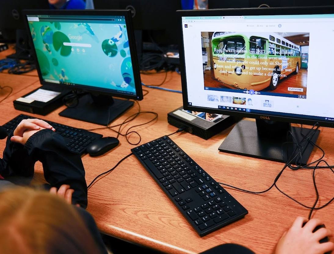 A student at a library computer builds a video slide about Rosa Parks, with the bus she rode on screen and the student’s own caption over it.