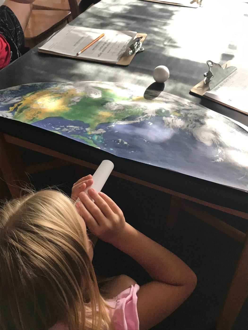 A student looks through a white viewing tube at a small moon model casting a shadow on a satellite image of Earth.