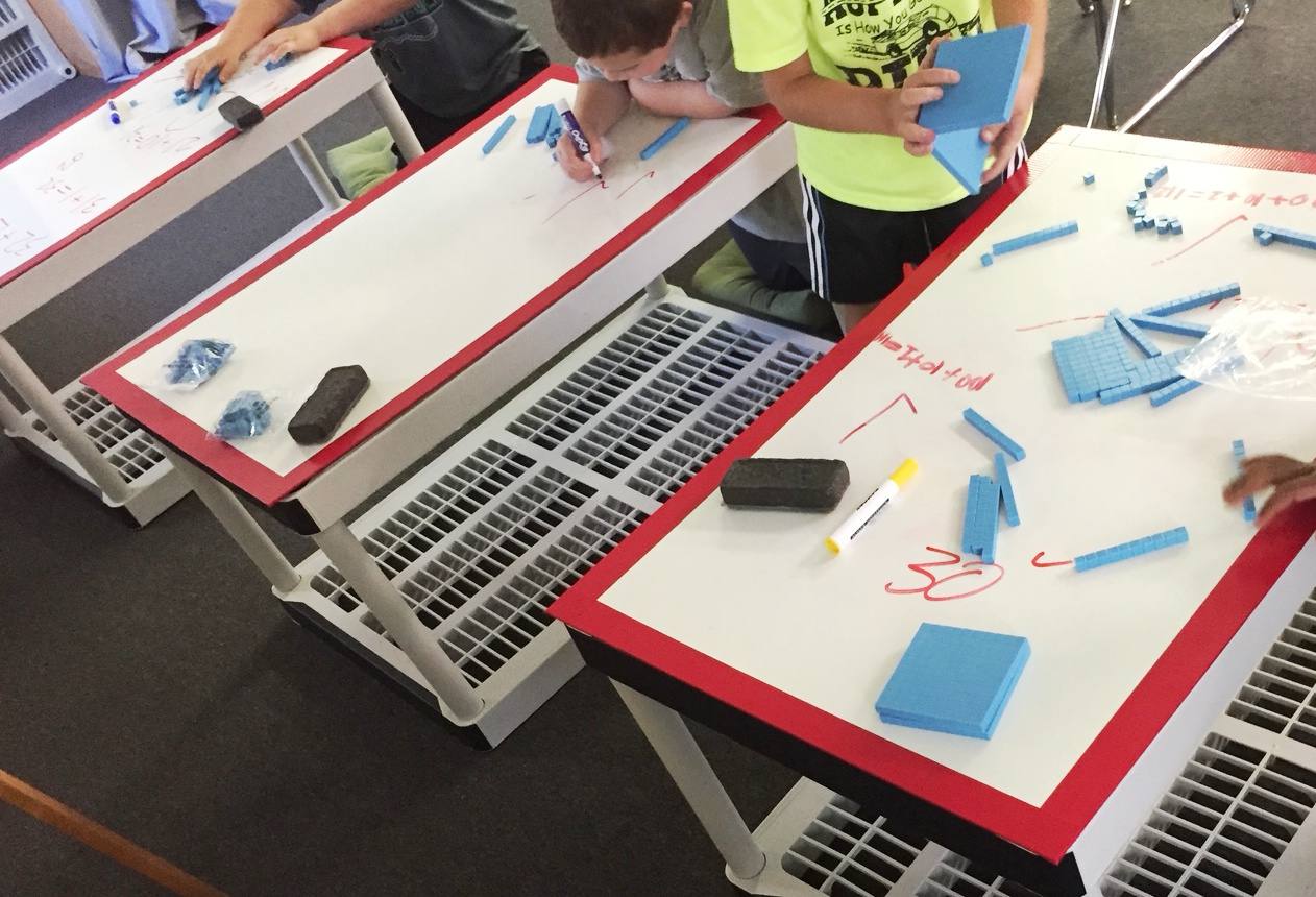 Students standing and kneeling at three red-edged whiteboard tables, building numbers with blue base-ten blocks and writing equations directly on the tabletops.